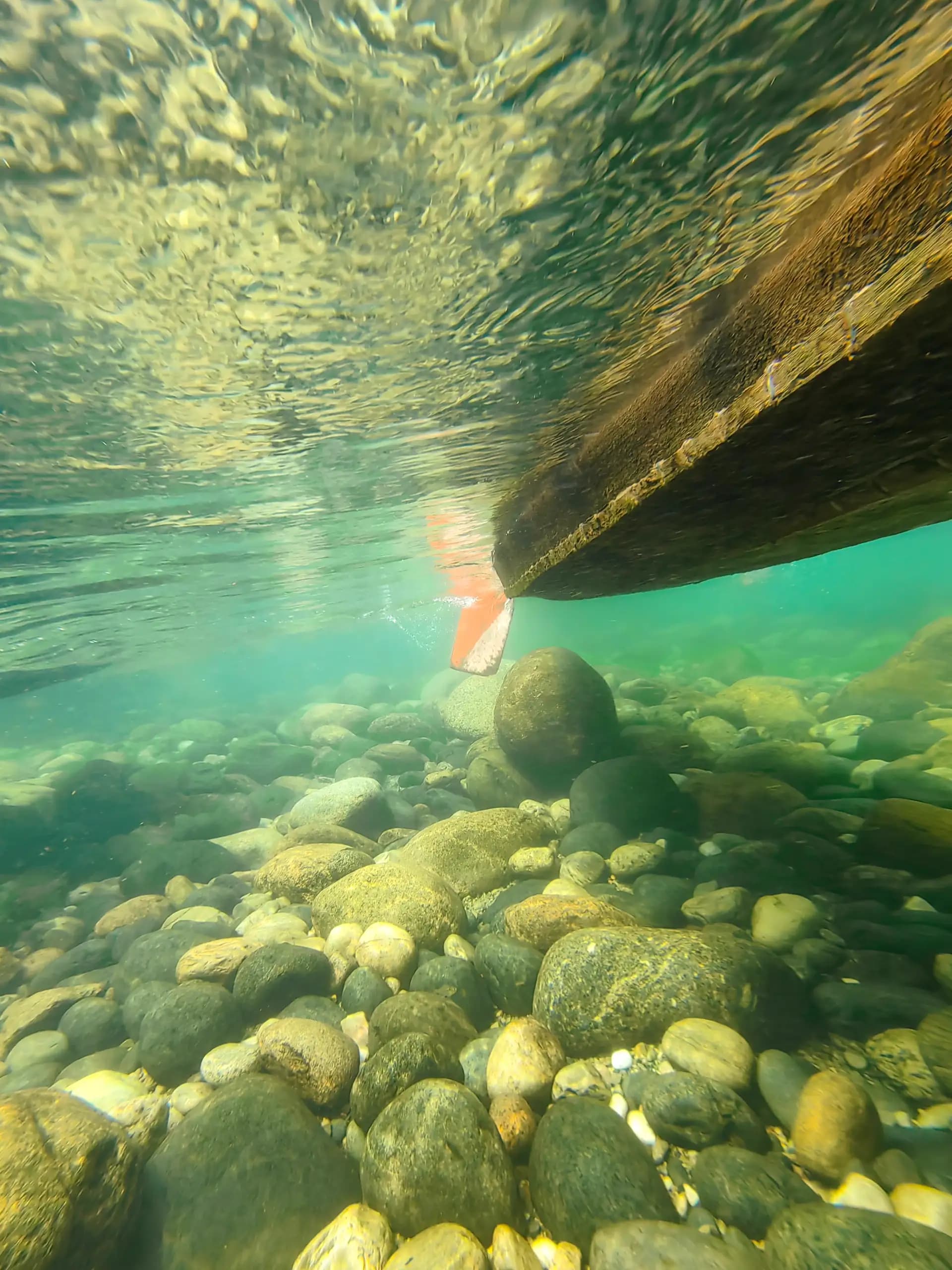 An underwater perspective beneath a drifting boat, revealing the quiet beauty of a crystal-clear riverbed hidden below the surface.